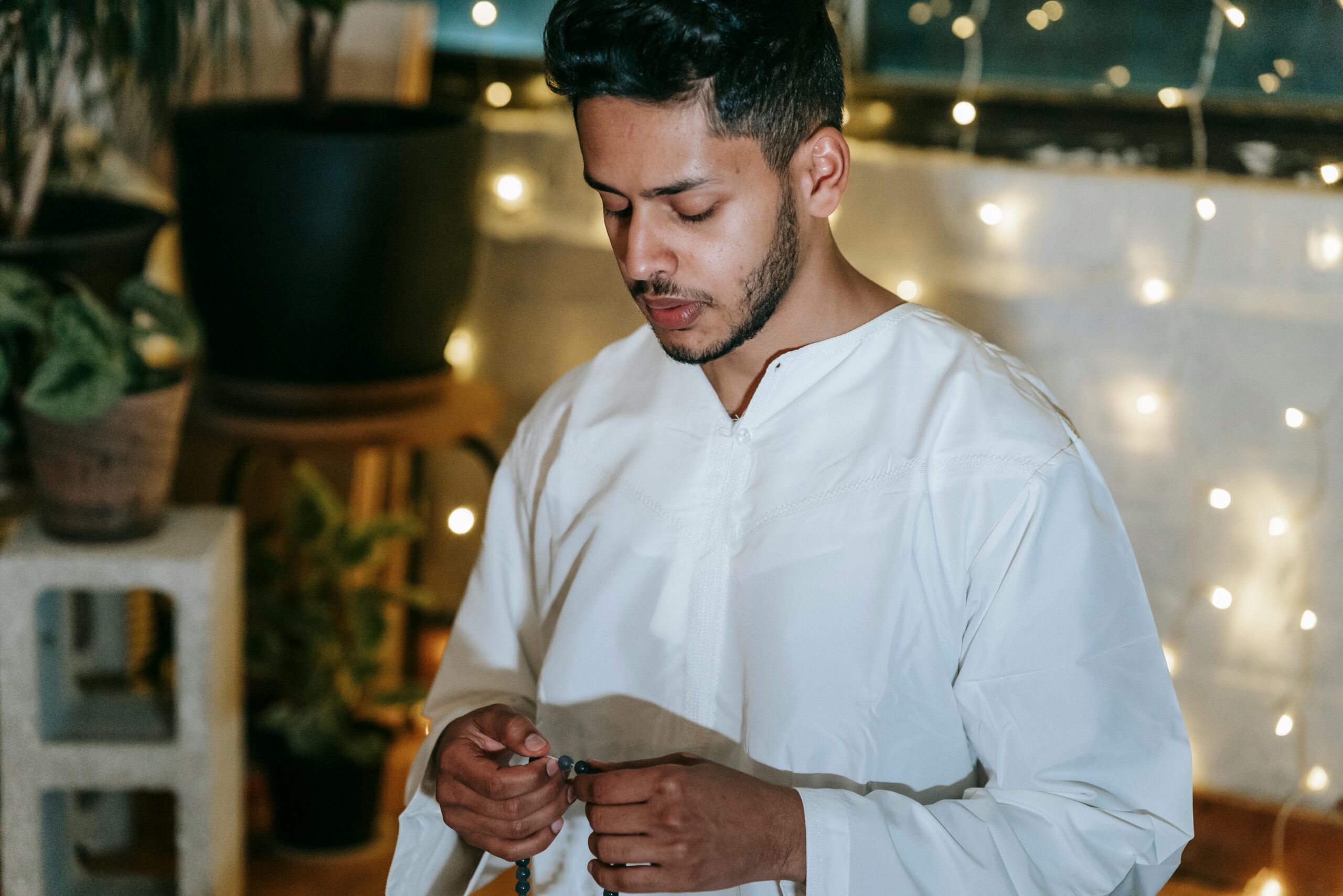 A man in traditional attire prays with beads indoors, with plants and string lights creating a calm atmosphere.