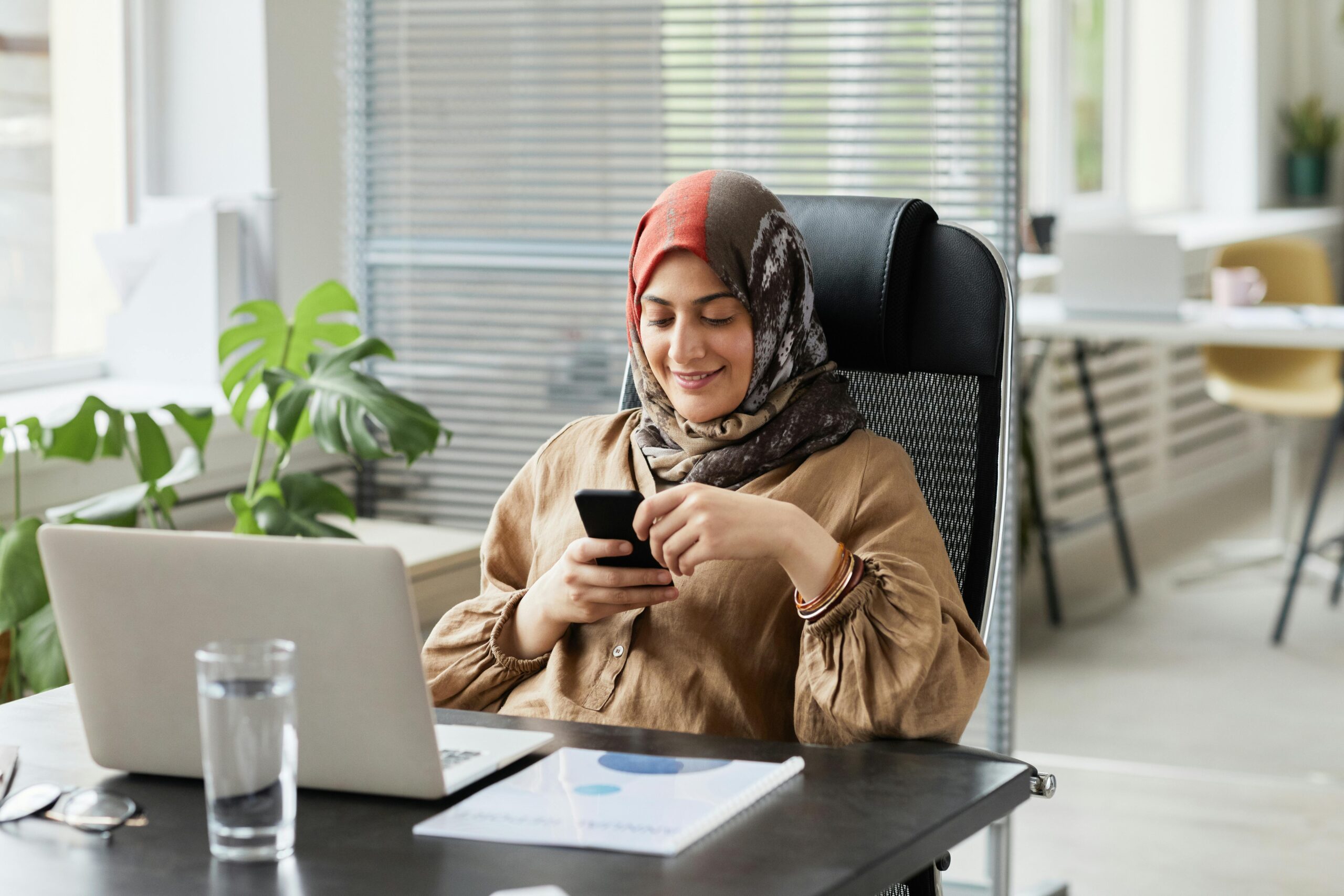 Confident businesswoman in hijab texting at her modern office desk with a laptop while smiling contentedly.